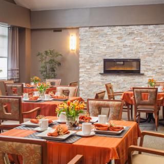 A warmly decorated dining room with tables set for a meal, featuring orange tablecloths and a modern fireplace.