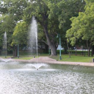 A tranquil park scene featuring fountains in a pond surrounded by lush trees.