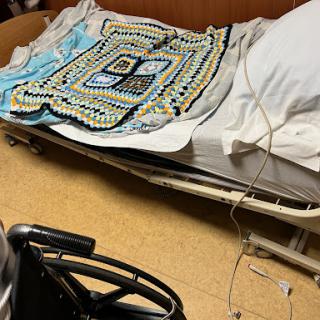 A hospital bed with a colorful blanket and a wheelchair nearby.