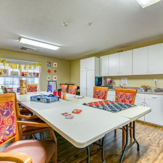 A brightly lit meeting room with a large table surrounded by chairs, adjacent to a kitchenette area.