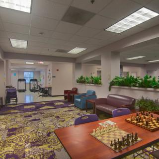 A cozy indoor seating area featuring a chess board on a table, surrounded by comfortable chairs and sofas, with decorative plants and bright lighting.