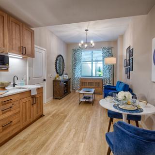 A modern open-concept living room and kitchen with wood flooring, featuring blue chairs and a small dining table.