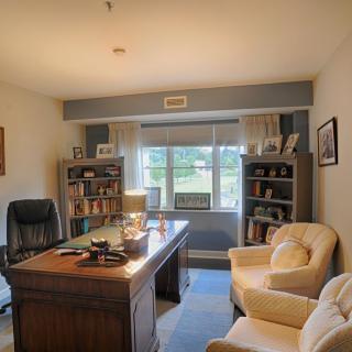 A cozy home office with a large desk, comfortable armchairs, and bookshelves under natural light from a window.