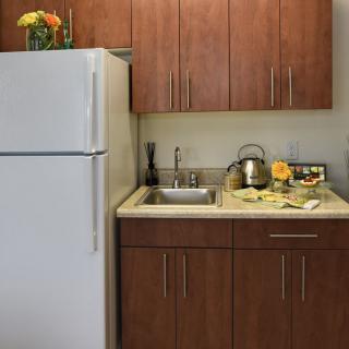 A small kitchen area with wooden cabinets, a refrigerator, and a countertop featuring a sink, kettle, and flowers.