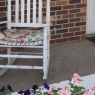 A white rocking chair sits on a porch with colorful flowers in the foreground and a brick wall in the background.