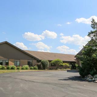 A wide view of a single-story building with an adjacent parking lot, surrounded by trees under a partly cloudy sky.