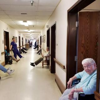 Elderly residents in wheelchairs line the hallway of a nursing home, accompanied by caretakers.