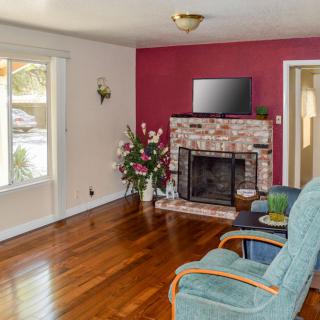 A cozy living room with hardwood floors, a brick fireplace, and a television mounted above. The room features a red accent wall, large window, armchair, and decorative plant.