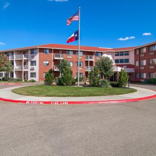 A three-story apartment complex with American and Texas flags flying in a well-landscaped roundabout on a sunny day.