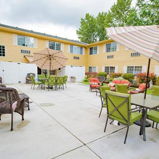 A spacious outdoor patio area with tables, chairs, and umbrellas, surrounded by a two-story yellow building.