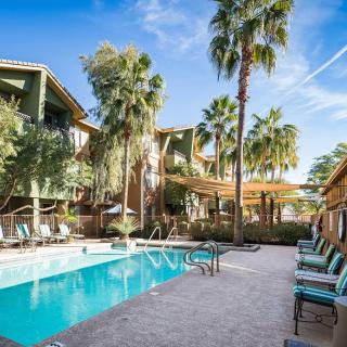 A tranquil apartment complex with a swimming pool surrounded by palm trees and outdoor seating on a sunny day.