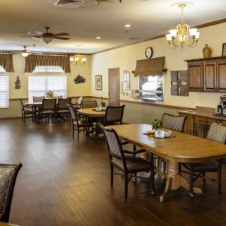 A cozy dining area with wooden tables and chairs, complemented by warm lighting and a kitchen in the background.
