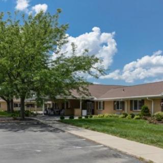 A single-story building surrounded by trees, with a parking lot and sidewalk under a partly cloudy sky.