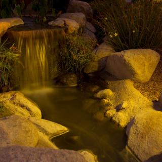 A serene nighttime garden waterfall illuminated by soft lighting, surrounded by rocks and lush greenery.