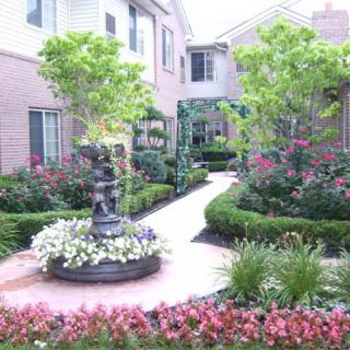 A beautifully landscaped garden with a walkway, surrounded by flowers and greenery, featuring a central fountain.