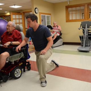 A healthcare professional assists a senior in a wheelchair with physical exercises in a rehabilitation center.