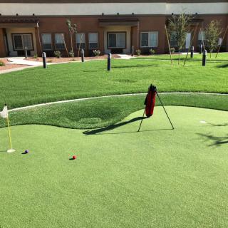 A small putting green with a golf bag and flagstick in front of a building.