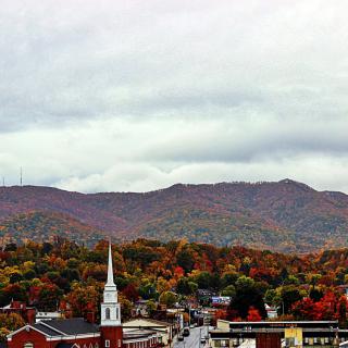 A scenic view of a town surrounded by colorful autumn foliage with mountains in the background under a cloudy sky.