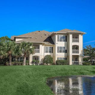 A modern apartment building with a pond and palm trees in the foreground under a clear blue sky.