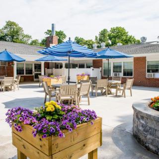 A sunny outdoor patio with tables, chairs, and blue umbrellas surrounded by flower planters.