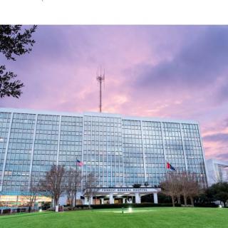 A large building with a glass facade at sunset, featuring a flag and surrounded by trees and a green lawn.