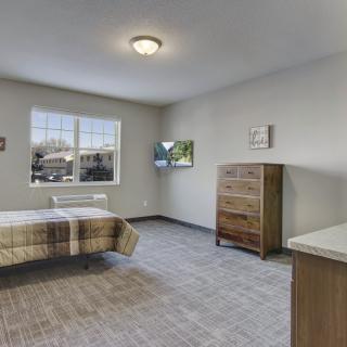 A minimalist bedroom with neutral tones, featuring a bed, dresser, and large window.