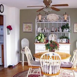 A cozy dining room with a round wooden table and white chairs, adorned with plants and a bookshelf in the background.