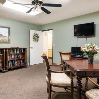 A cozy living room featuring a wooden table with chairs, a bookshelf filled with books, a wall-mounted television, and decorative flowers on the table.