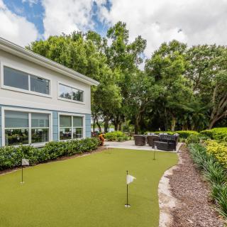 A backyard featuring a putting green and a patio area surrounded by lush greenery.