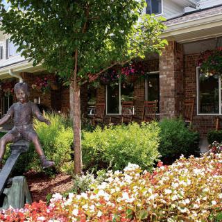 A charming garden scene featuring a sculpture amidst vibrant flowers and a brick building in the background.