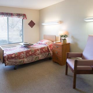 A simple hospital room featuring a bed, chair, and natural light from the window.