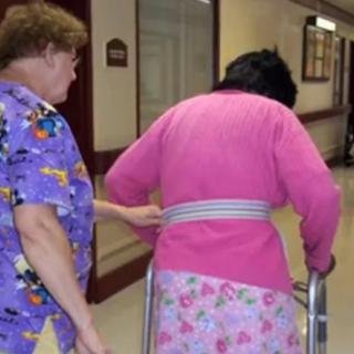 A healthcare worker assists an elderly person using a walker in a hallway.