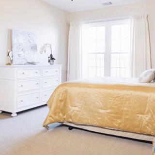 A bright, well-lit bedroom featuring a gold bedspread and white furniture.