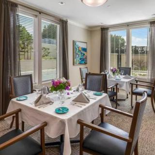 A well-decorated dining room with a neatly set table, surrounded by chairs, featuring large windows and elegant curtains.