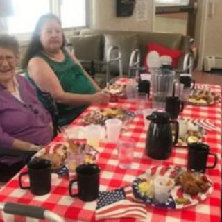 A group of people sitting at a table with food on a red checkered tablecloth, decorated with American flags.