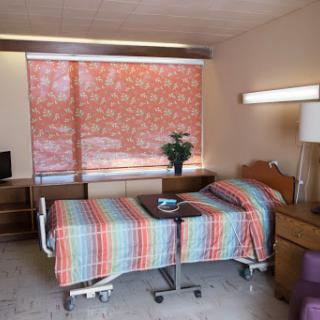 A well-lit hospital patient room featuring a single bed, desk, and a window adorned with patterned blinds.