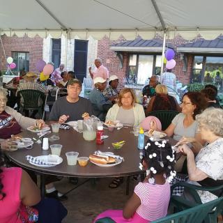 A group of people enjoying a meal together under a tent, with balloons in the background.