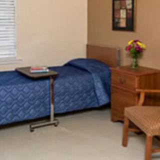 A simple bedroom with a single bed, a blue bedspread, a wooden nightstand with flowers, and a chair.