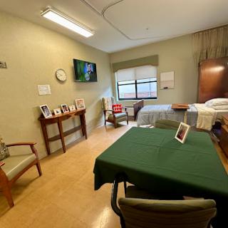 A well-lit hospital room featuring a bed, a table with chairs, and personal photographs.