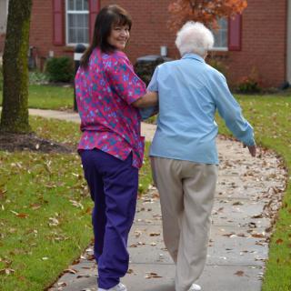 A caregiver assists an elderly person during a walk on a leaf-strewn path.