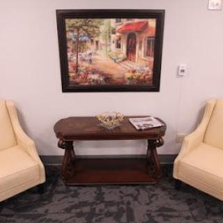 A cozy waiting area with two beige chairs, a wooden table, and a landscape painting on the wall.