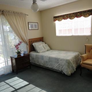 A cozy bedroom featuring a single bed, a nightstand with flowers, and a comfortable chair by the window.