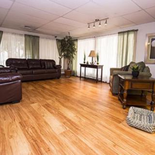 A spacious living room with wooden flooring, featuring a brown leather sofa, side tables, and large windows with curtains.