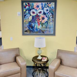 A cozy interior space featuring two armchairs, a side table with a lamp, and a vibrant floral painting on the wall.