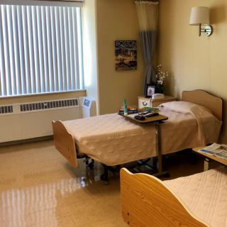 A hospital room with two beds, a window with blinds, and a bedside table.
