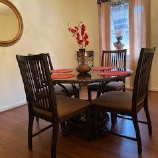 A cozy dining area with a round wooden table and four chairs, adorned with a vase of red flowers. A round mirror decorates the wall, complementing the warm tones of the room.