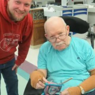 An elderly man in a wheelchair holding an item, with another man standing beside him.