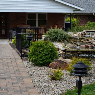 A serene pathway leading to a charming home, featuring a tranquil water feature and lush greenery.