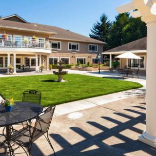 A sunny patio area with outdoor seating, surrounded by a well-maintained garden and a two-story building in the background.