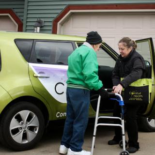 A caregiver assists an elderly person with a walker as they prepare to enter a vehicle.